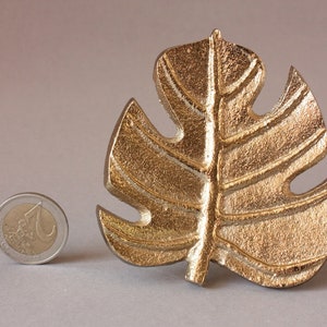 May include: A gold-colored metal leaf with a detailed design, resembling a monstera leaf. The leaf is about the size of a US quarter.