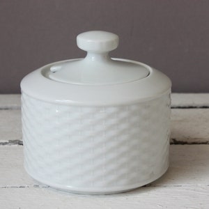 Op de afbeelding: Een witte keramische suikerpot met een deksel en een kleine knop. De kom heeft een getextureerd, geweven patroon rond de cilindrische behuizing. Het deksel heeft een kleine opening voor een lepel. De suikerpot staat op een wit houten oppervlak.