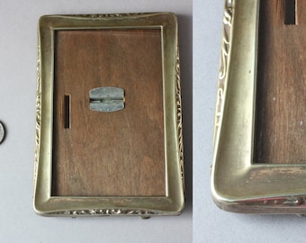 May include: Rectangular antique picture frame with a gold-colored metal frame and a wooden backing. The frame has decorative scrollwork and a hinged metal clasp. A 2 Euro coin is next to the frame for size comparison.