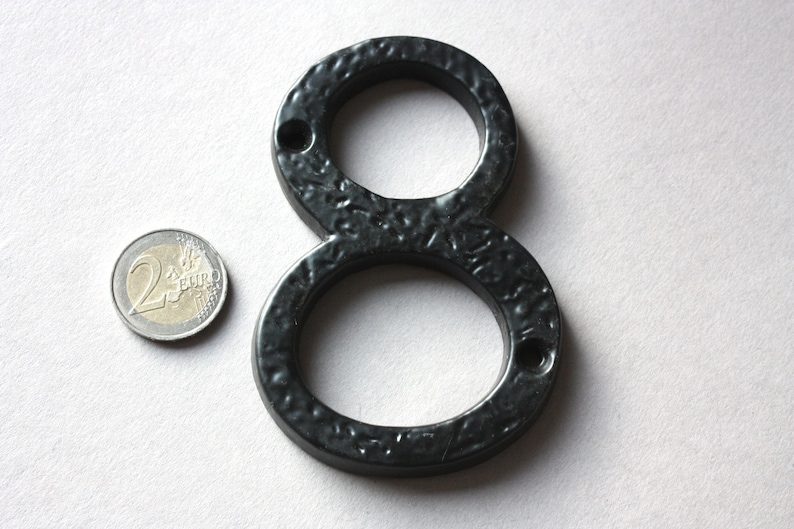 K&ouml;nnte beinhalten: Schwarze Hausnummer acht aus Metall. Die Nummer besteht aus einem einzigen St&uuml;ck Metall und hat eine strukturierte Oberfl&auml;che. Die Nummer ist etwa so gro&szlig; wie eine Zwei-Euro-M&uuml;nze.
