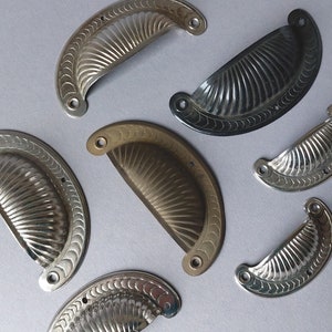 Könnte beinhalten: Eine Sammlung von acht dekorativen Schranktürgriffen in verschiedenen Ausführungen, darunter Silber, Messing und Schwarz. Die Griffe sind alle halbmondförmig mit einem gewellten Design.