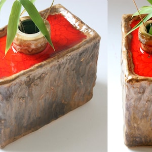 Könnte beinhalten: Eine rechteckige Keramikvase mit einer rauen, strukturierten Außenseite und einem glatten, rot glasierten Innenraum. Die Vase hat eine kleinere, runde Öffnung in der Mitte für Blumen oder Pflanzen. Die Vase ist mit Wasser und einem einzigen Bambusstock gefüllt.