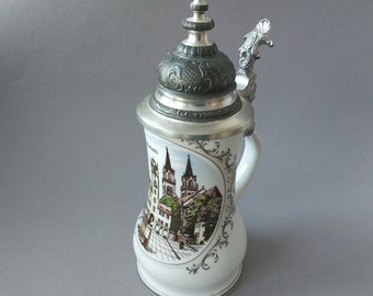 May include: A white ceramic beer stein with a pewter lid and handle. The stein features a detailed illustration of a German town with buildings and a church.