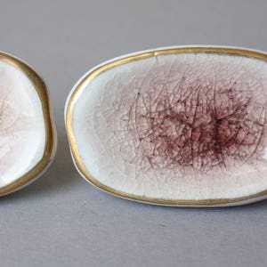 以下が含まれることがあります： 金色の縁取りとピンクのひび割れ釉薬を施した、白いセラミック製の楕円形の皿が2つあります。