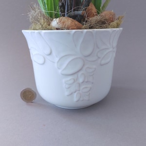 Pode incluir: Um vaso de flores de cerâmica branca com desenhos florais em relevo. O vaso tem uma forma larga e curva e é preenchido com terra, musgo seco e elementos decorativos. Uma moeda está ao lado do vaso para escala.