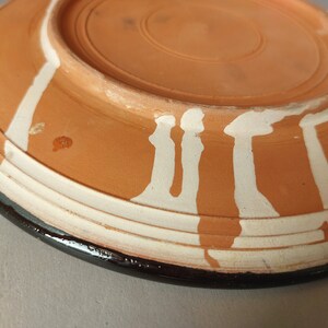 May include: A terracotta plate with a white glaze design. The plate has a black rim and a raised edge.