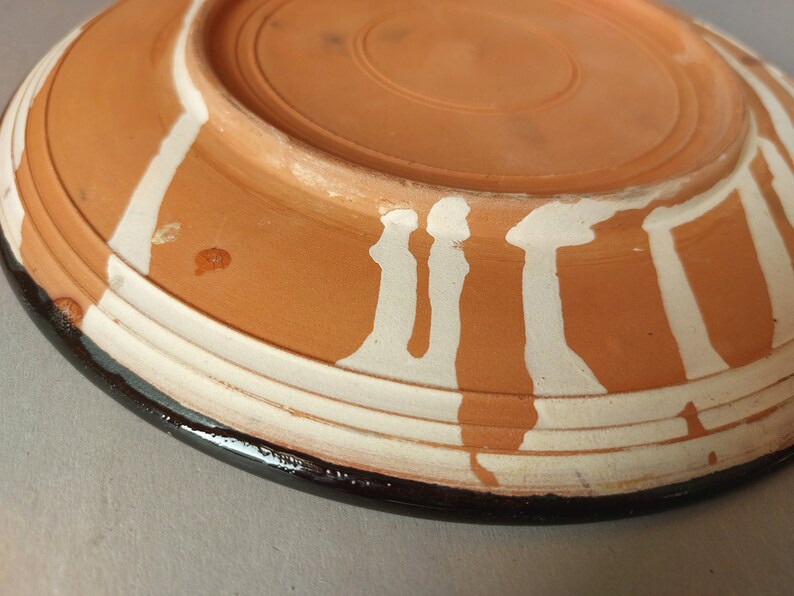 May include: A terracotta plate with a white glaze design. The plate has a black rim and a raised edge.
