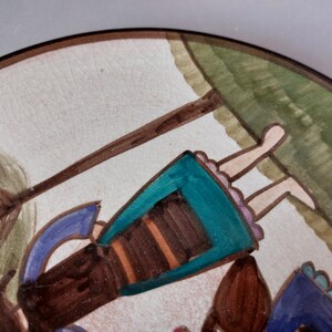 May include: A ceramic plate with a hand-painted design of two girls swinging on a rope swing. The girls are wearing colorful dresses and have brown hair. The background is a green field with a brown tree.