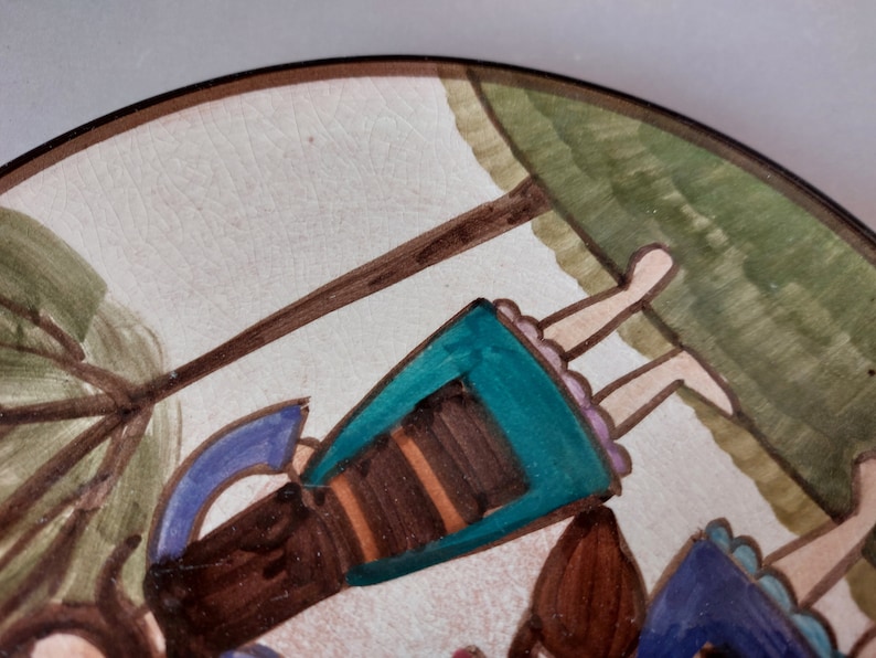 May include: A ceramic plate with a hand-painted design of two girls swinging on a rope swing. The girls are wearing colorful dresses and have brown hair. The background is a green field with a brown tree.