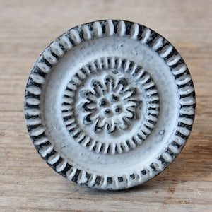 Könnte beinhalten: Weißer Keramik-Knopf mit einem schwarz-weißen Blumendesign. Der Knopf hat einen erhabenen, strukturierten Rand.