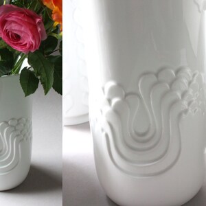 May include: Two white ceramic vases with a raised, abstract design. The vase on the left is holding a bouquet of pink and orange flowers.