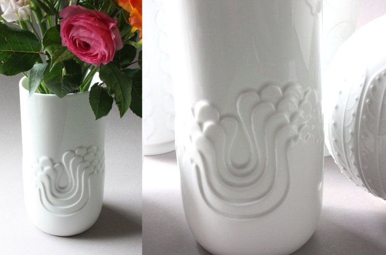 May include: Two white ceramic vases with a raised, abstract design. The vase on the left is holding a bouquet of pink and orange flowers.