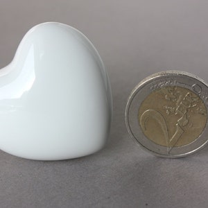 Puede incluir: Un adorno de cerámica blanca en forma de corazón junto a una moneda de 2 euros para comparar el tamaño.