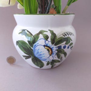 May include: White ceramic flower pot with a hand-painted blue and green floral design. The pot has a rounded shape and a green rim. A coin is next to the pot. The pot is filled with artificial flowers.