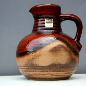 May include: A brown and beige ceramic pitcher with a wide, round body and a handle. The pitcher has a brown glaze with a black stripe and a beige stripe. The pitcher has a label that says "BAY Keramik".