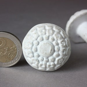 Może przedstawiać: Dwa białe ceramiczne gałki do szafek z fakturowanym, wypukłym wzorem. Jeden gałka jest okrągły z okrągłym wzorem, drugi jest bardziej tradycyjnym gałką.