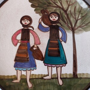 May include: A ceramic plate with a hand-painted design of two women in traditional clothing carrying jugs. The women are standing in front of a tree with green leaves and a brown trunk. The plate has a white background with brown and green accents.