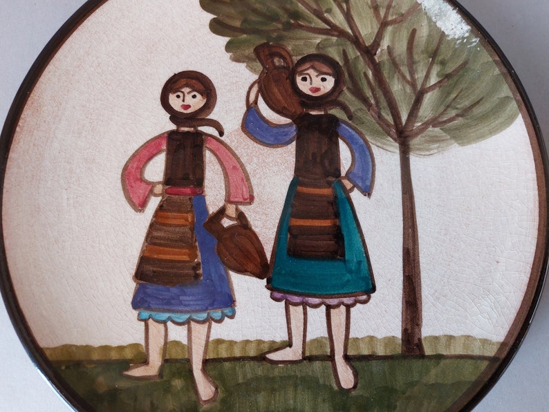 May include: A ceramic plate with a hand-painted design of two women in traditional clothing carrying jugs. The women are standing in front of a tree with green leaves and a brown trunk. The plate has a white background with brown and green accents.