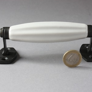 Może przedstawiać: Biały ceramiczny klamka do drzwi z czarnymi metalowymi akcentami. Klamka ma owalny kształt i żebrowany wzór. Klamka jest zamontowana na dwóch czarnych metalowych płytkach.