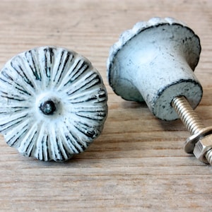 Könnte beinhalten: Zwei weiße Metall-Schrankknöpfe mit einem dekorativen, erhabenen Sonnenstrahlen-Design. Ein Knopf ist von der Seite zu sehen, wodurch die Schraube und die Mutter sichtbar werden.