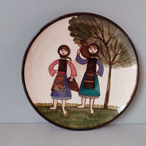 May include: A round ceramic plate with a painted scene of two women in traditional clothing carrying jugs, standing in front of a green tree. The plate has a brown rim.