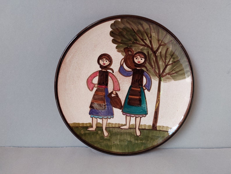 May include: A round ceramic plate with a painted scene of two women in traditional clothing carrying jugs, standing in front of a green tree. The plate has a brown rim.