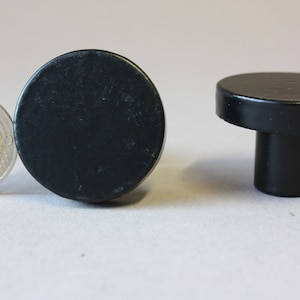 Könnte beinhalten: Zwei schwarze runde Schrankknöpfe. Ein Knopf ist flach und der andere hat einen kurzen, zylindrischen Stiel.