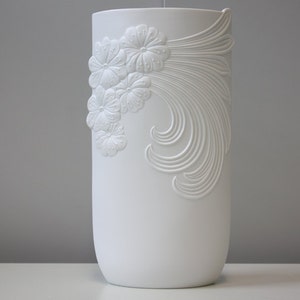 May include: White ceramic vase with an embossed floral design. The vase is tall and cylindrical with a smooth, white finish.