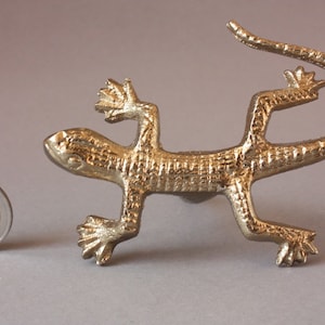 May include: A gold-coloured metal lizard figurine with a textured surface. The lizard is posed with its tail curled up and its head tilted to the side.