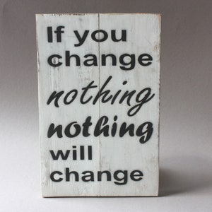 May include: A white wooden sign with black text. The sign displays the message: "If you change nothing, nothing will change." The wood has a distressed finish, with visible grain and a slightly worn look.