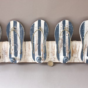 Op de afbeelding: Een houten wandbevestigde kapstok in de vorm van vijf slippers. De slippers zijn wit geverfd met zwarte strepen en hebben een verweerde look. Elke slipper heeft een metalen haak om items aan op te hangen.