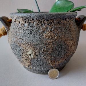 Op de afbeelding: Een donkergrijze keramische pot met een ruw, getextureerd oppervlak. De pot heeft twee touwhandvatten en een bruine, gevlekte glazuur. Een euro-muntstuk ligt voor de pot voor schaal.