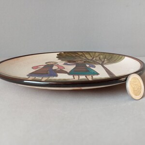 May include: A white ceramic plate with a brown rim features a hand-painted design of two figures in traditional clothing standing under a green tree.