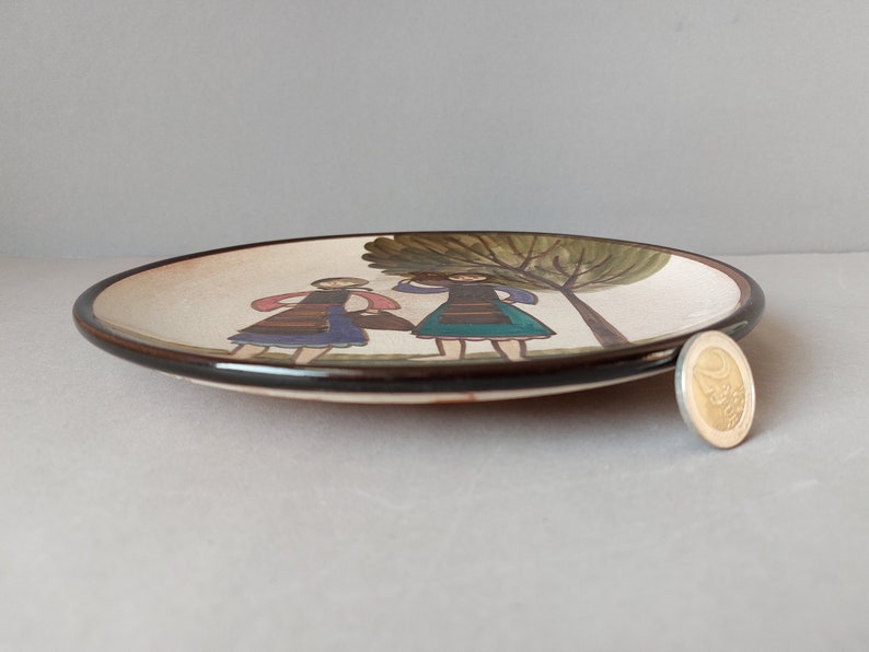 May include: A white ceramic plate with a brown rim features a hand-painted design of two figures in traditional clothing standing under a green tree.