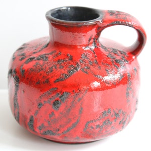 May include: Red ceramic vase with a black glaze and a textured surface. The vase has a wide, round body and a narrow neck with a handle.