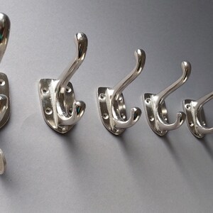 Könnte beinhalten: Fünf silberne Metallhaken mit einem Doppelhaken-Design, die an einer grauen Wand montiert sind.