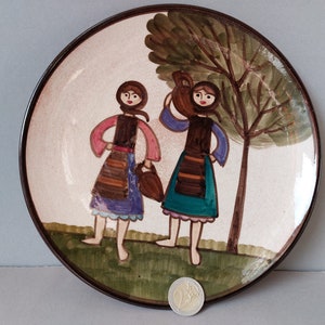 May include: A ceramic plate with a brown rim and a white background. Two women in colorful dresses are painted on the plate, carrying buckets. A green tree is painted behind them.