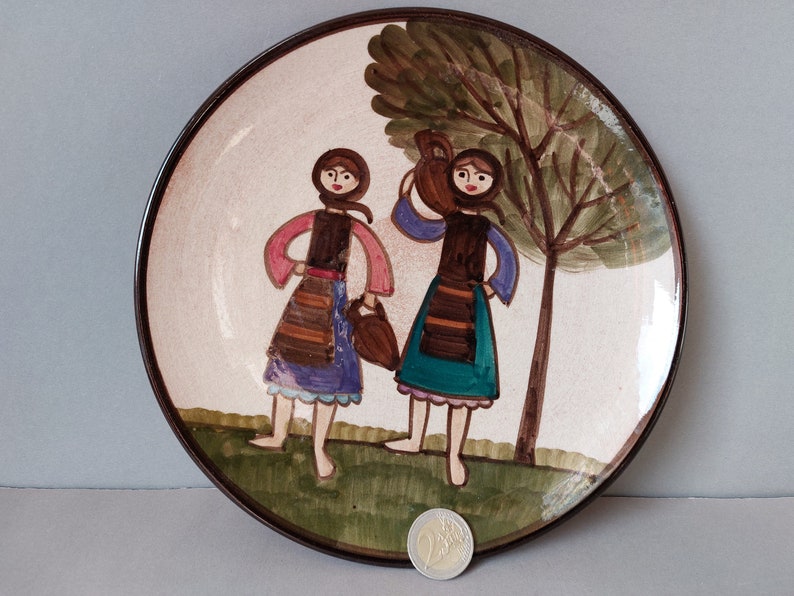 May include: A ceramic plate with a brown rim and a white background. Two women in colorful dresses are painted on the plate, carrying buckets. A green tree is painted behind them.