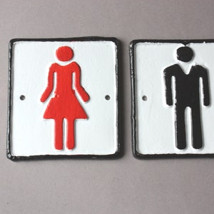 Könnte beinhalten: Zwei weiße Metallschildchen mit schwarzen Rändern. Das Schild links hat ein rotes weibliches Symbol und das Schild rechts ein schwarzes männliches Symbol.