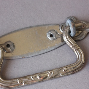 May include: A silver-toned metal drawer pull with an ornate design. The pull has a rectangular plate with a curved handle. The handle is decorated with a floral pattern.