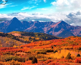 Puede incluir: Una cordillera de montañas con un cielo azul y nubes blancas. En primer plano, un bosque de árboles con hojas rojas, naranjas y amarillas.