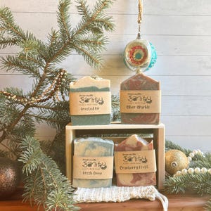 May include: Four bars of goat's milk soap in a wooden crate. The soaps are decorated with swirls of green, blue, and red. The labels on the soaps read "Preserving My Sanity Goat's Milk Soap" with the names "Frosted Fir", "Silver Sparkle", "Fresh Snow", and "Cranberry Frost".