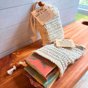 May include: Handmade soap bars in a natural woven bag with a drawstring closure. The soap bars are various colors, including pink, blue, and brown. The tag reads "Odds & Ends". The bag is sitting on a wooden surface.