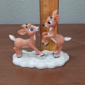 May include: A figurine of two reindeer on a snowy base. The reindeer are brown with white details and black hooves. One reindeer is on its hind legs, the other on all fours. The figurine is approximately 10 cm tall.