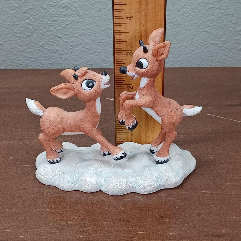 May include: A figurine of two reindeer on a snowy base. The reindeer are brown with white details and black hooves. One reindeer is on its hind legs, the other on all fours. The figurine is approximately 10 cm tall.
