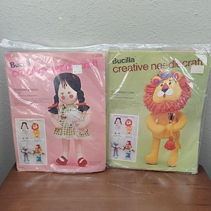 May include: Two Bucilla creative needlecraft kits. One kit features a doll of Dorothy and Toto, the other a Cowardly Lion. The Dorothy doll has black pigtails and a green and white dress. The lion is yellow with a red mane and a tassel.