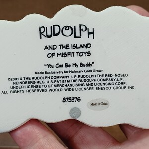 May include: A white figurine featuring the text "RUDOLPH AND THE ISLAND OF MISFIT TOYS" and the phrase "You Can Be My Buddy". The figurine is made exclusively for Hallmark Gold Grown. The text includes copyright and trademark details.