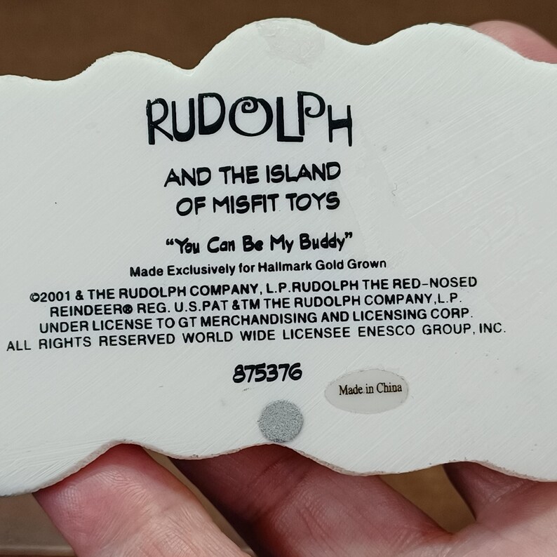 May include: A white figurine featuring the text "RUDOLPH AND THE ISLAND OF MISFIT TOYS" and the phrase "You Can Be My Buddy". The figurine is made exclusively for Hallmark Gold Grown. The text includes copyright and trademark details.