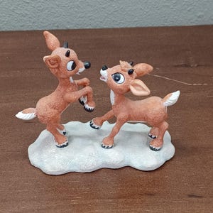 May include: A ceramic figurine of two reindeer in a playful pose. The reindeer are brown with white accents and black hooves, standing on a snow-covered base. The figurine has a cartoonish style and is likely a Christmas decoration.