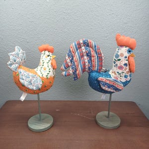 May include: Two decorative rooster and hen figurines. The rooster has a blue body and a patterned tail with red, white, and blue stripes. The hen has an orange and white body with a floral pattern. Both have orange combs and are mounted on green stands.